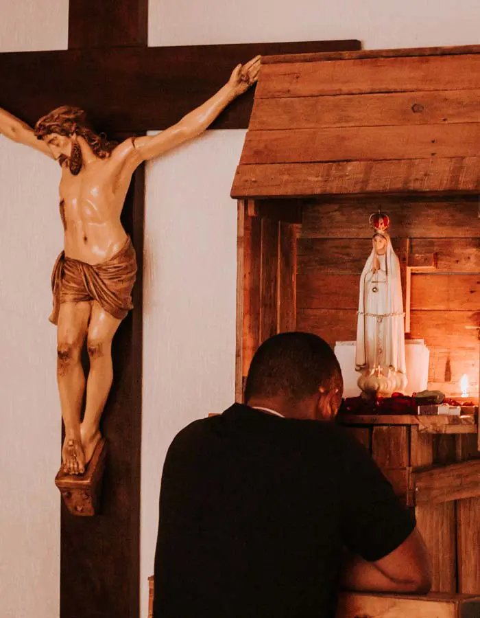 um homem rezando em frente uma imagem de Maria Santíssima e ao lado uma grande cruz com Jesus crucificado